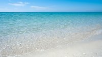 Pristine tropical beach with crystal clear turquoise water gently lapping white sand shore under bright blue sky with soft clouds for vacation travel.
