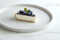 Slice of creamy cheesecake topped with fresh blueberries and mint leaf on white ceramic plate against marble background. Minimalist food photography.