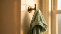 Green towel hanging on brass hook in warm sunlit bathroom with white subway tiles and wooden window frame creating cozy morning atmosphere.