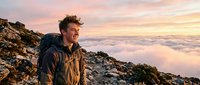 Young man with brown hair and light complexion standing on rocky mountain summit at sunrise, gazing at dramatic cloud-filled valley below. Wearing dark jacket. Perfect for adventure, travel, and lifestyle content.