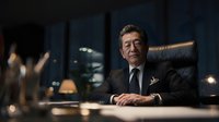 Senior Asian businessman in elegant suit sitting at conference table in modern office boardroom during evening meeting with city lights background.