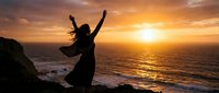 Silhouetted woman with arms raised celebrating freedom on rocky coastal cliff at golden sunset over ocean waves.