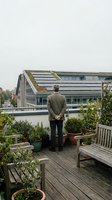 Mature man in business casual attire standing on wooden rooftop terrace with potted plants, overlooking modern sustainable architecture with green roof and solar panels.