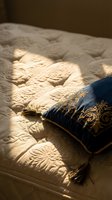 Luxurious quilted bedding with ornate navy pillow featuring gold embroidered details and tassel trim, bathed in warm natural sunlight for bedroom interior design and home decor projects.