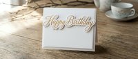 Elegant white greeting card with gold Happy Birthday text on rustic wooden table with teacup and dried branches for celebration design.