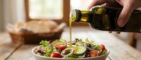 Young adult hand pouring golden olive oil over fresh vegetable salad with tomatoes, cucumbers, and greens in white bowl on wooden table for cooking and food preparation.