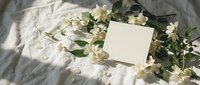 Blank white card surrounded by fresh white jasmine flowers and green leaves on textured linen fabric with natural sunlight shadows, suitable for wedding invitations, greeting cards, and stationery design.