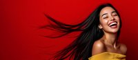 Young Asian woman with flowing black hair laughing joyfully against vibrant red background, wearing yellow off-shoulder top, expressing genuine happiness and freedom.