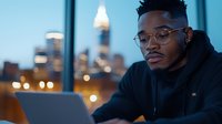Young African American man working on laptop computer in modern office during evening hours with city skyline background for business and technology concepts.