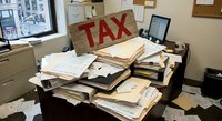 Cluttered office desk overflowing with tax documents, papers and files during busy accounting season with computer and window view.