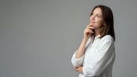 Thoughtful young woman in white shirt looking up with hand on chin in contemplative pose against neutral background for business and lifestyle concepts.