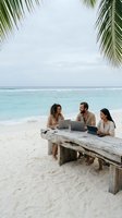 Remote work team meeting on tropical beach with laptops under palm trees discussing business strategy on wooden table with ocean view.
