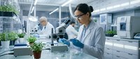 Asian female scientist in white lab coat conducting research with microscope in modern laboratory facility with colleague working in background.