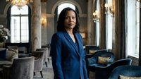 Professional African American businesswoman in elegant blue suit standing confidently in luxury hotel lobby with marble columns and sophisticated interior design.
