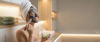 Young woman with towel on head applying facial mask in modern bathroom holding cucumber slice for skincare routine and spa treatment at home.