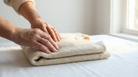 Senior man with light skin tone folding beige linen towel on white surface near window, domestic household chore and laundry care routine.