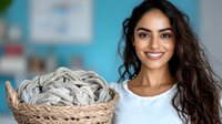Young woman with long dark wavy hair and bright smile holding wicker basket with gray wool yarn balls and knitting supplies against blurred blue background.