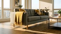 Charcoal gray upholstered sofa with wooden frame and gold accents in bright living room with large windows, natural light, and warm earth tone decor for interior design and home furnishing projects.