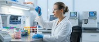 Female scientist in white lab coat and protective gloves conducting research with test tubes in modern laboratory setting with medical equipment and analysis machines.