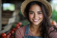 Young Hispanic female farmer in straw hat and overalls smiling at farmers market with fresh produce and tomatoes in background, representing agriculture and local food.