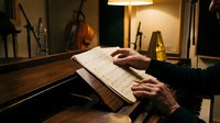 Musician reading sheet music at piano with warm studio lighting, hands positioned over keyboard during practice session or composition work.