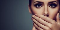 Young woman covering mouth with hands, expressing shock, fear or silence against dark background. Dramatic portrait with intense gaze.
