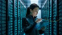Female IT professional with dark hair and South Asian ethnicity working in a server room, holding a fiber optic cable while examining network infrastructure with digital displays.