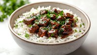 Tender braised beef chunks in rich brown sauce served over fluffy white rice, garnished with fresh chopped parsley in ceramic bowl on light background.
