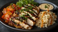 Grilled chicken breast slices with fresh herbs, served alongside colorful tomato salad, steamed rice and creamy sauce on dark plate, close up food photography.