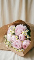 Delicate pink peony bouquet wrapped in brown kraft paper with white chamomile flowers on neutral fabric background for romantic occasions and floral gifts.