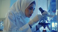 Young female scientist of Middle Eastern descent wearing white hijab and lab coat, conducting microscopic research in modern laboratory setting with scientific equipment and glassware.