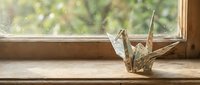 Origami paper crane sits on wooden windowsill with soft natural light streaming through glass, creating peaceful zen atmosphere for meditation and mindfulness.