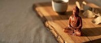 Wooden Buddha statue in meditation pose on rustic wooden tray with tea cup and natural elements creating peaceful zen atmosphere for mindfulness and relaxation.