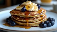 Stack of fluffy pancakes topped with fresh banana slices, ripe blueberries and drizzled with golden maple syrup on white plate, close up view of breakfast.