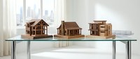 Wooden architectural model houses displayed on glass table in modern office with city skyline view through windows for real estate presentation.