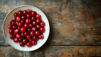 Fresh ripe cranberries on white ceramic plate against rustic wooden background with copy space, top view composition for food photography.