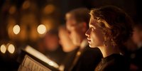 Young woman reading sheet music in warm candlelight during evening choir performance with blurred singers in background creating atmospheric concert setting.