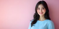 Young Asian woman with long dark hair wearing light blue blouse smiling warmly against pink background, professional portrait with copy space for business or lifestyle content.