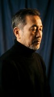 Mature Hispanic man in black turtleneck sweater looking thoughtfully to side with dramatic lighting against dark blue curtain background for portrait photography.