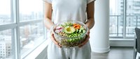 Woman in casual gray shirt holding fresh salad bowl with vegetables and greens in modern high-rise apartment with city skyline view, healthy eating and nutrition concept.