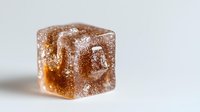 Brown sugar cube covered in crystallized sugar particles on white background, macro close-up view showing texture and sparkle details in soft lighting.