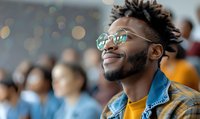 Young African American man with stylish sunglasses and denim jacket smiling confidently at outdoor event, expressing joy and optimism against blurred bokeh background.