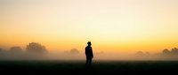 Silhouetted male figure standing alone in misty field at sunrise, contemplating nature and solitude during golden hour for meditation and lifestyle concepts.