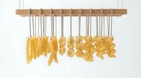 Assorted dried pasta hanging from wooden rod on strings against white background, showcasing various shapes and textures for culinary and food styling purposes.