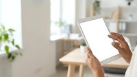 Hands holding tablet with blank white screen in bright modern home office with natural light, plants, and wooden furniture for app mockup and digital technology concepts.