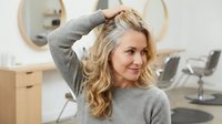 Middle-aged blonde Caucasian woman touching her hair in modern salon interior with styling chairs and mirrors in background.