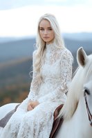 Young Caucasian woman with long platinum blonde hair wearing white lace wedding dress sitting on white horse against mountain landscape background in soft evening light.