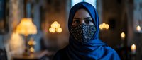 Young Muslim woman wearing blue hijab and decorative face mask in warm indoor setting with bokeh lights creating cozy atmospheric background for lifestyle content.