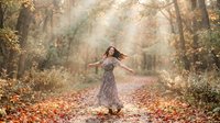 Woman with long dark hair, wearing a floral patterned dress, spinning with arms outstretched on a forest path covered in autumn leaves with golden sunlight filtering through trees.
