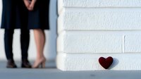 Romantic couple standing near white brick wall with small red heart decoration, legs and feet visible only. Minimalist love concept for valentines and dating.